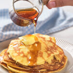 A hand pours maple syrup from a small pitcher onto a stack of pancake.