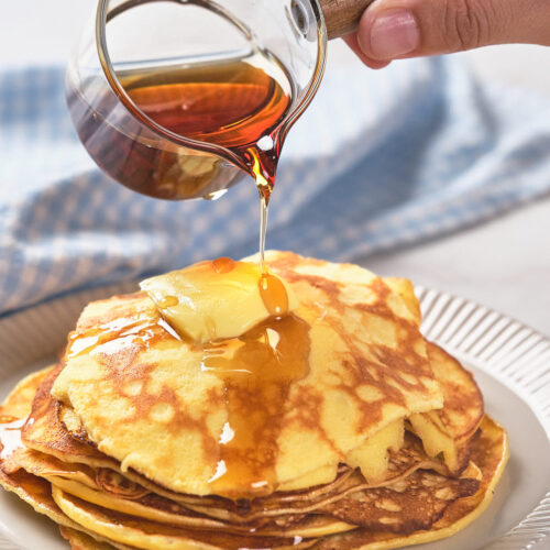 A hand pours maple syrup from a small pitcher onto a stack of pancake.