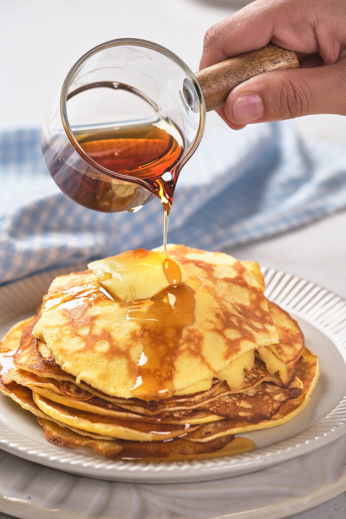 A hand pours maple syrup from a small pitcher onto a stack of pancake.