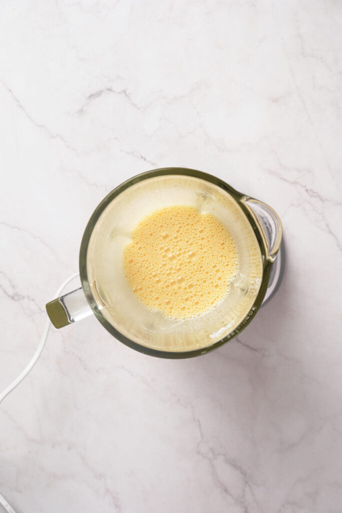 A top-down view of a blender containing a frothy yellow mixture on a white marble countertop.