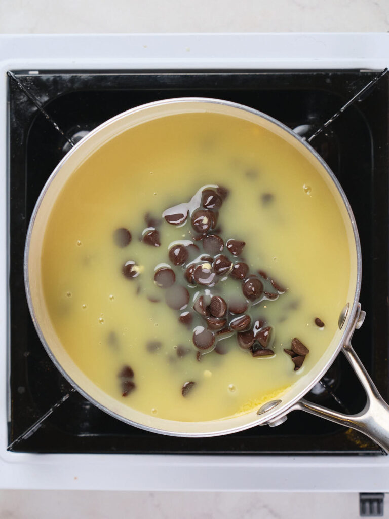 A saucepan on a stovetop contains condensed milk with chocolate chips starting to melt.