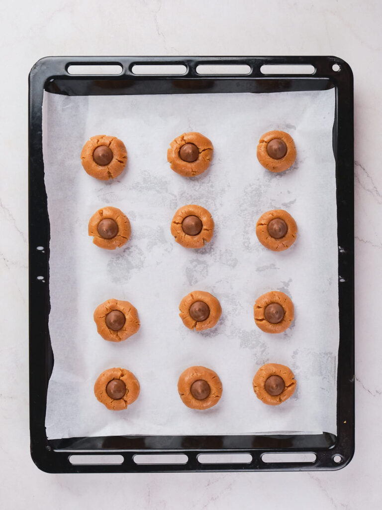 A baking tray lined with parchment paper holds twelve balls of peanut butter blossoms dough, each topped with a chocolate piece.