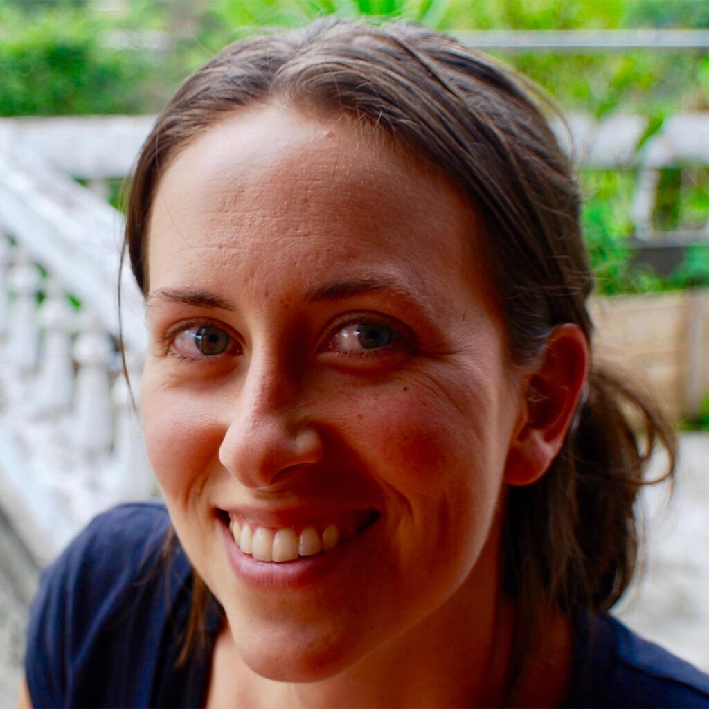 A woman with dark hair tied back, wearing a dark shirt, smiles at the camera with greenery in the background.