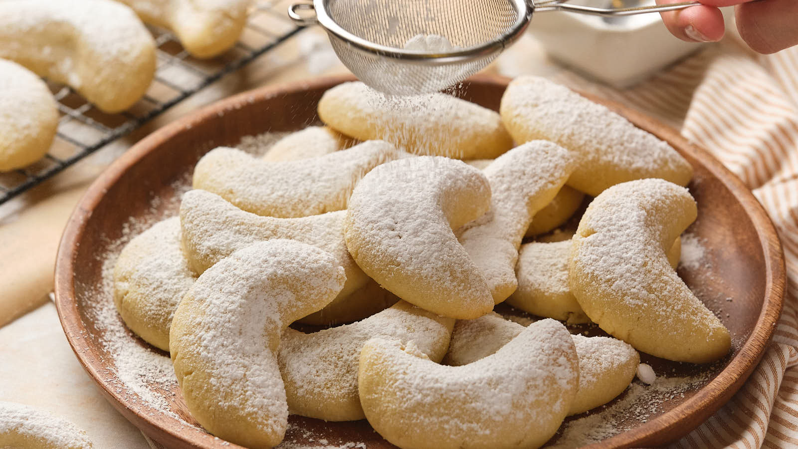 A plate of almond crescent cookies is being dusted with powdered sugar.