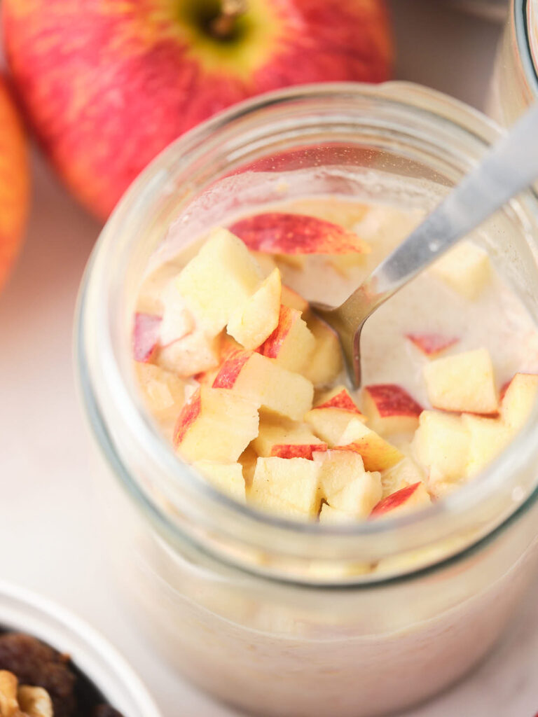 A jar filled with overnight oats topped with diced apples and a spoon inside.