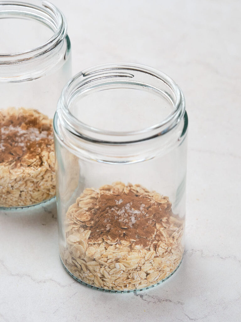 Two glass jars containing oats, cinnamon, and a sprinkling of salt on a white surface.