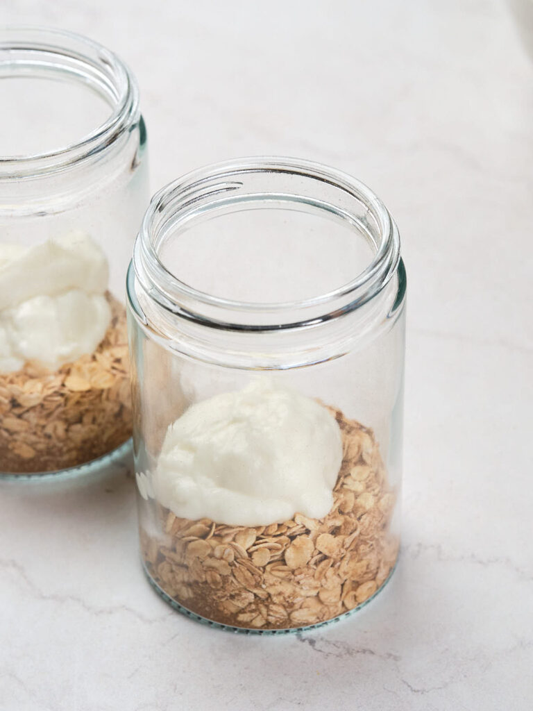 Two glass jars on a white surface, each containing a layer of dry oats and topped with a dollop of yogurt.