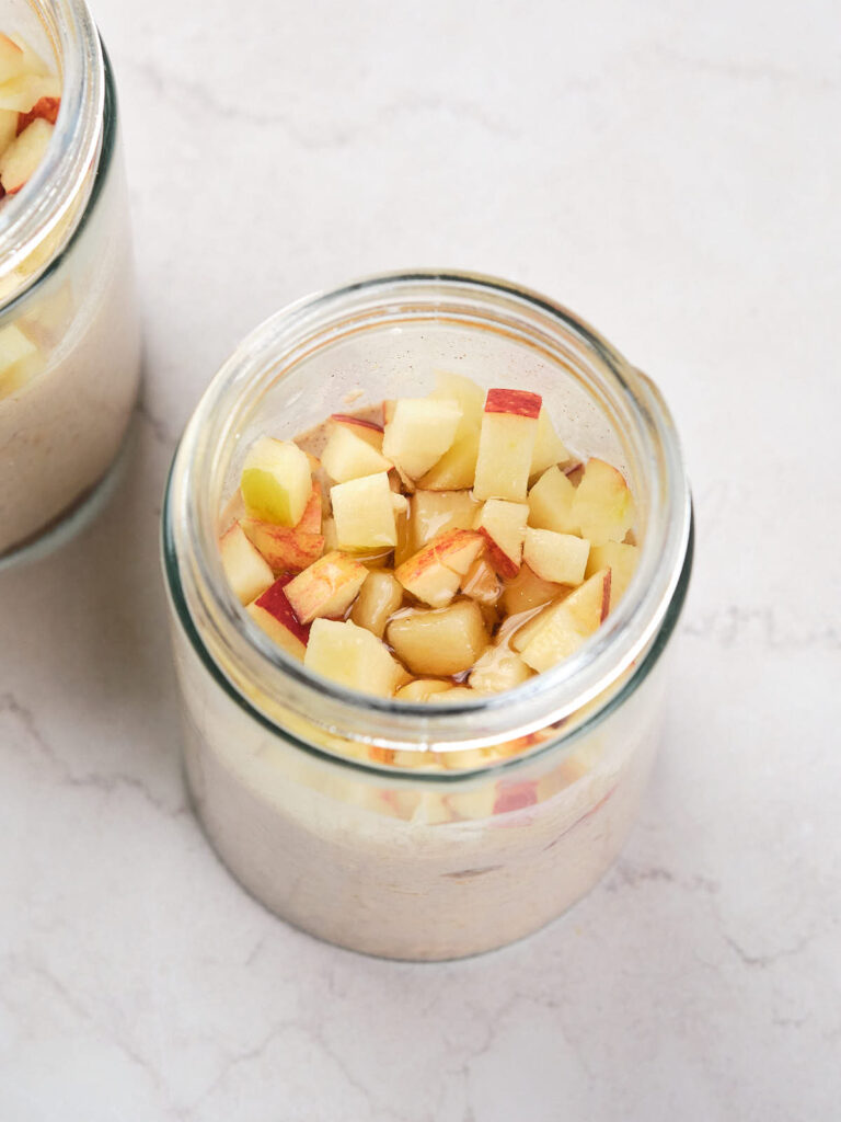 A glass jar filled with cinnamon overnight oats mixture, topped with small cubes of apples.