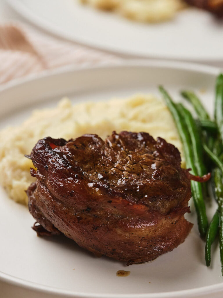 A plate featuring a bacon-wrapped fillet mignon, a serving of mashed potatoes, and a side of green beans.