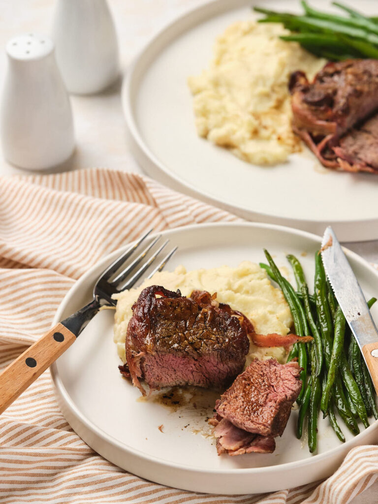 A plate with a serving of bacon wrapped fillet mignon, mashed potatoes, and green beans.