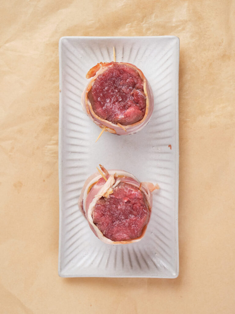 Two uncooked bacon-wrapped fillet mignons are placed on a rectangular white ceramic dish.