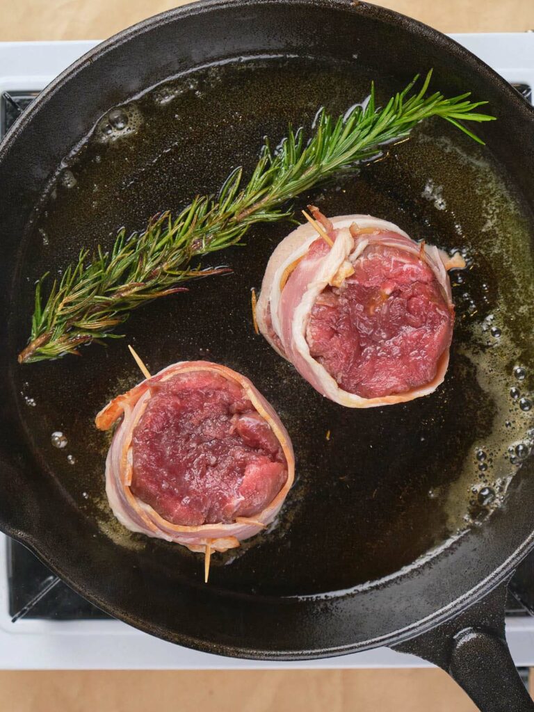 Two bacon-wrapped fillet mignon steaks and a sprig of rosemary are cooking in a black cast iron skillet.