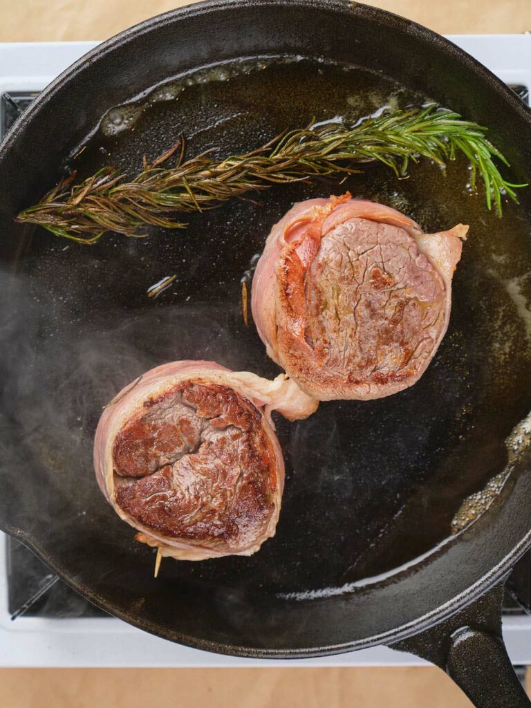 Two bacon-wrapped fillet mignon and a sprig of rosemary are being cooked in a black cast iron pan on a stove.