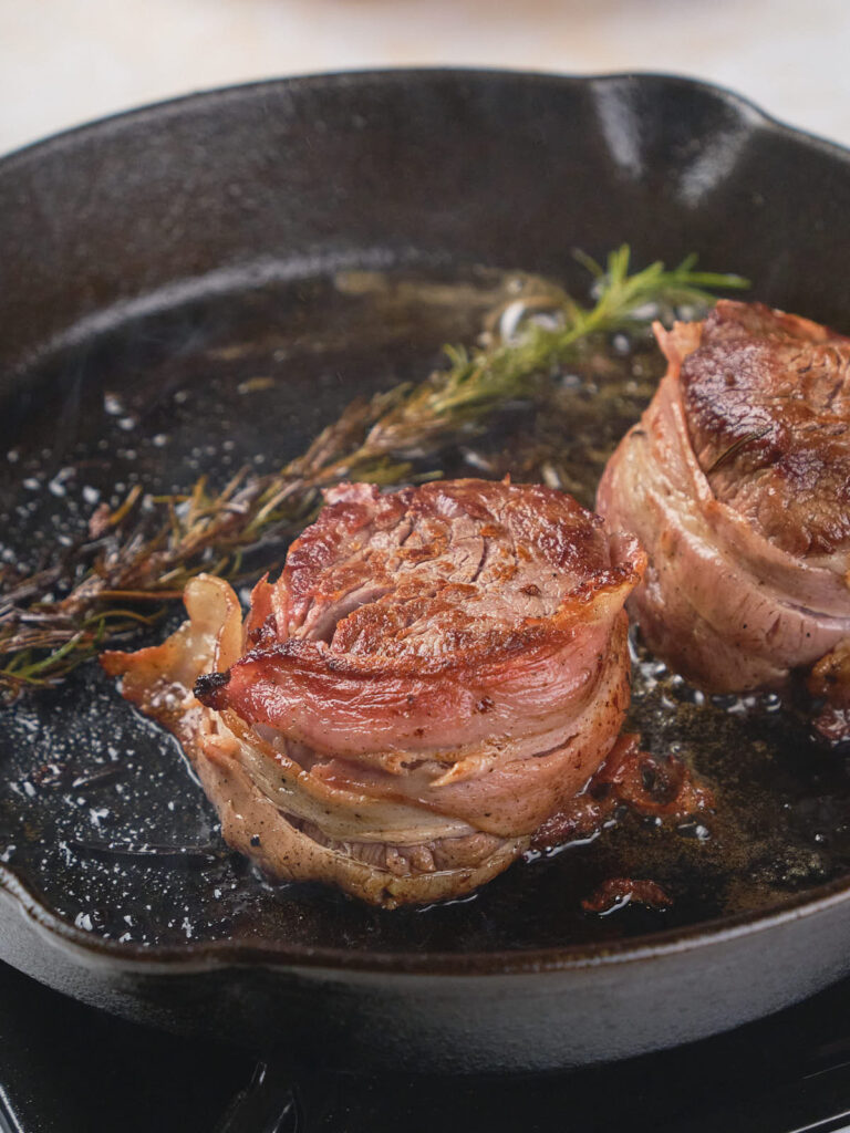 Two bacon-wrapped fillet mignon cooking in a black cast iron skillet, accompanied by a sprig of rosemary.