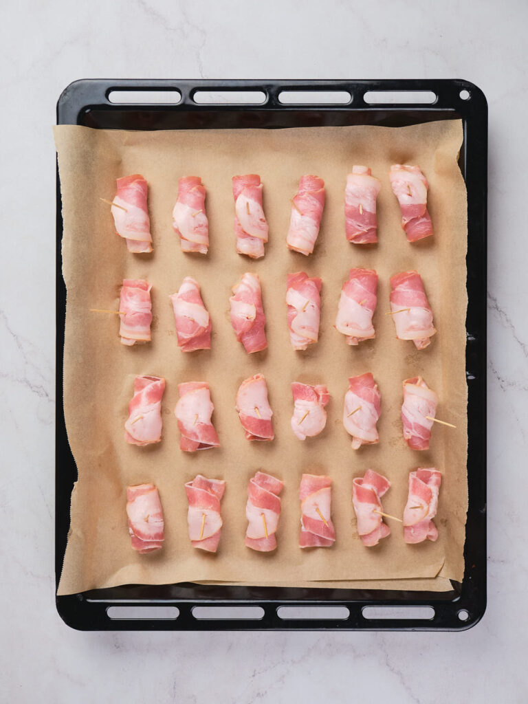 A baking tray lined with parchment paper holds multiple pieces of bacon-wrapped pineapple.