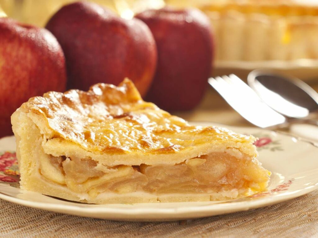 A slice of apple pie is served on a plate with a fork and spoon in the background.