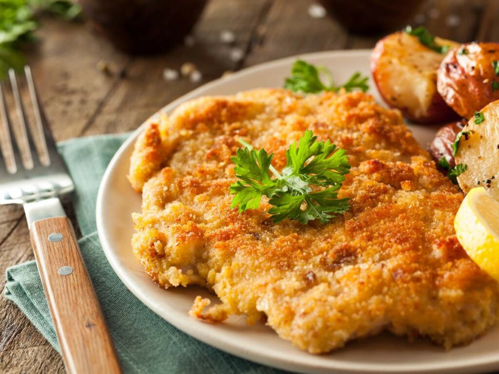 Chicken cutlet topped with parsley is served with roasted potatoes and a lemon wedge on a white plate.