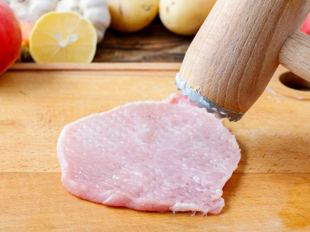 A wooden meat tenderizer pounds a raw piece of meat on a wooden cutting board.