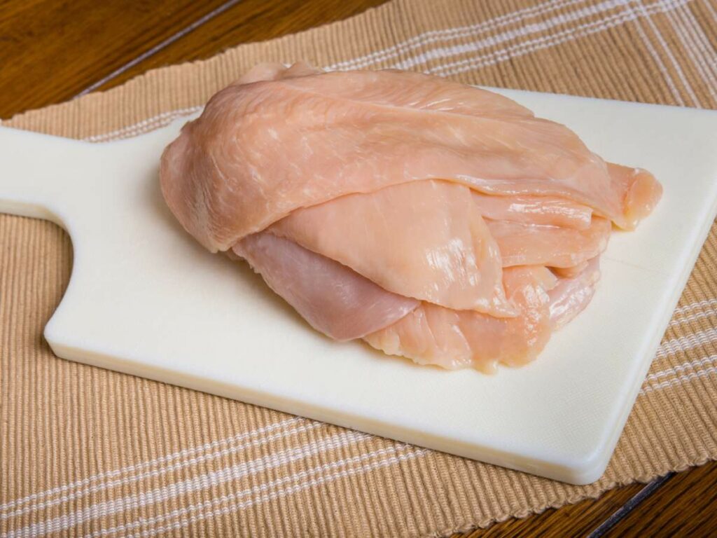 Raw chicken breast fillets stacked on a white cutting board.