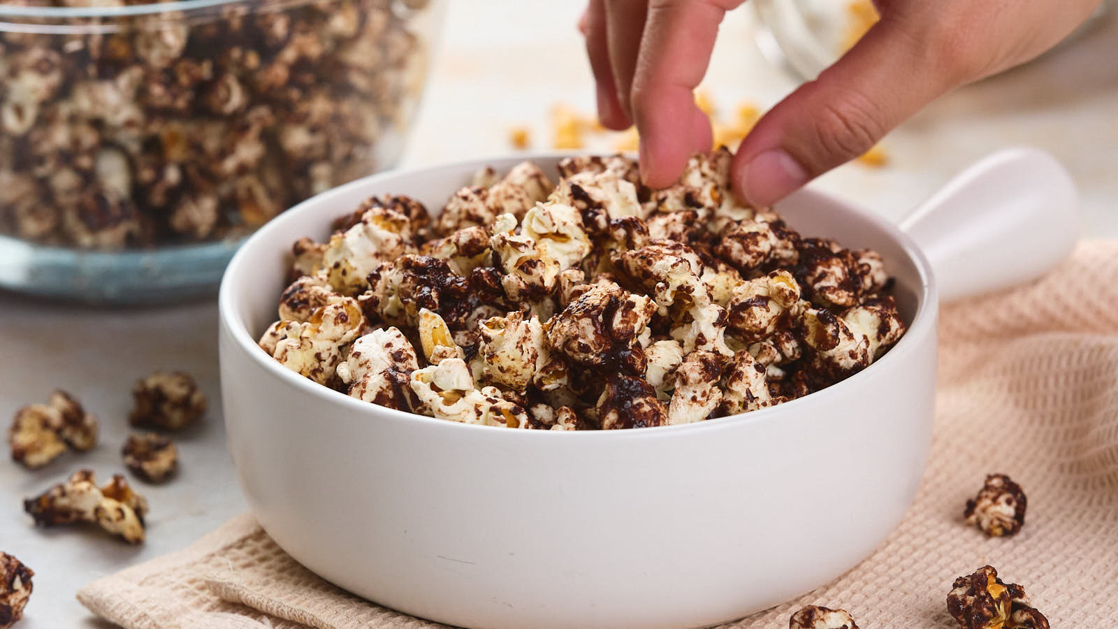 A hand reaches for a piece of chocolate-coated popcorn in a white bowl.