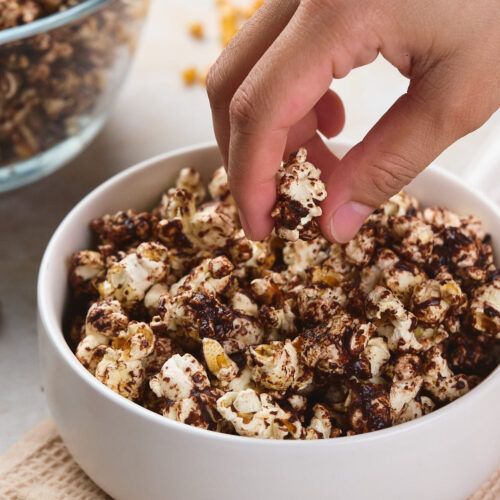 A hand is picking a piece of chocolate-covered popcorn from a white bowl.