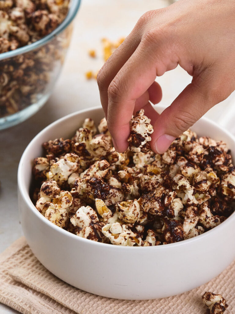 A hand is picking a piece of chocolate-covered popcorn from a white bowl.