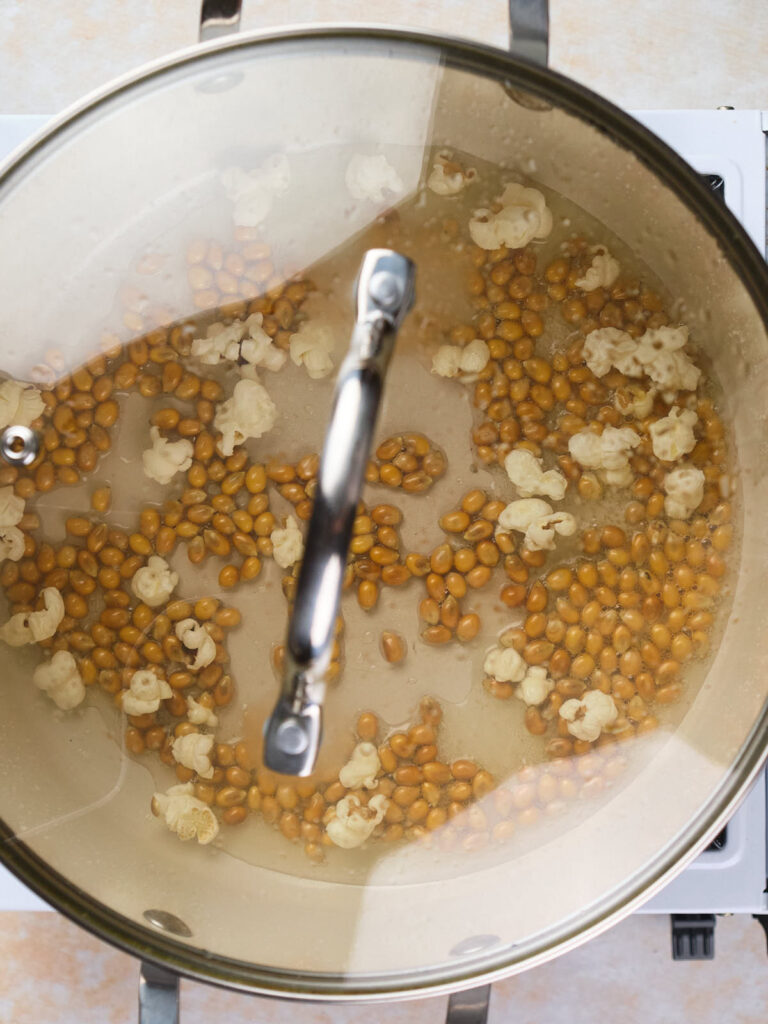 A glass-covered pot on a stovetop containing partially popped popcorn kernels in oil.