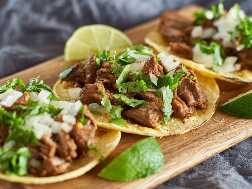 Three beef tacos garnished with chopped onions and cilantro on a wooden board.