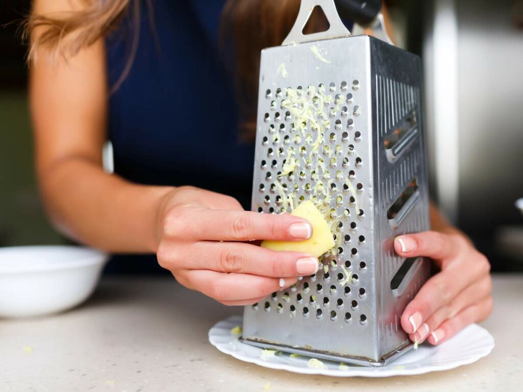 Close-up of a person grating a piece of cheese using a box grater over a white plate on a kitchen counter.