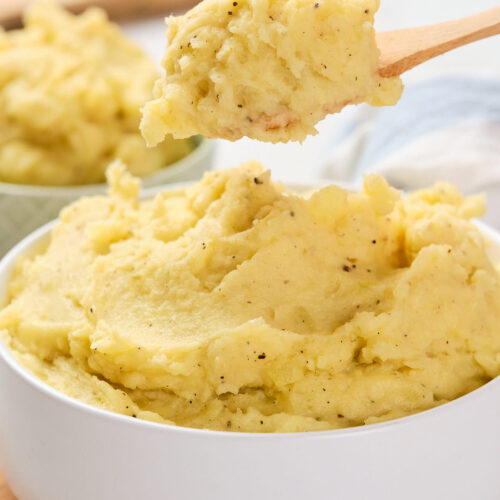 A white bowl filled with garlic mashed potatoes, with a wooden spoon lifting a portion from the bowl.