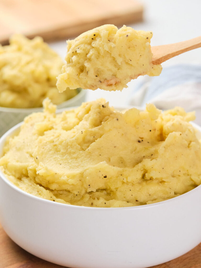 A white bowl filled with garlic mashed potatoes, with a wooden spoon lifting a portion from the bowl.