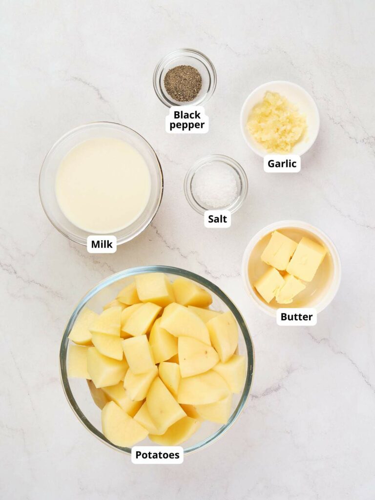 A bowl of chopped potatoes, dishes of black pepper, garlic, salt, and butter, and a small bowl of milk.