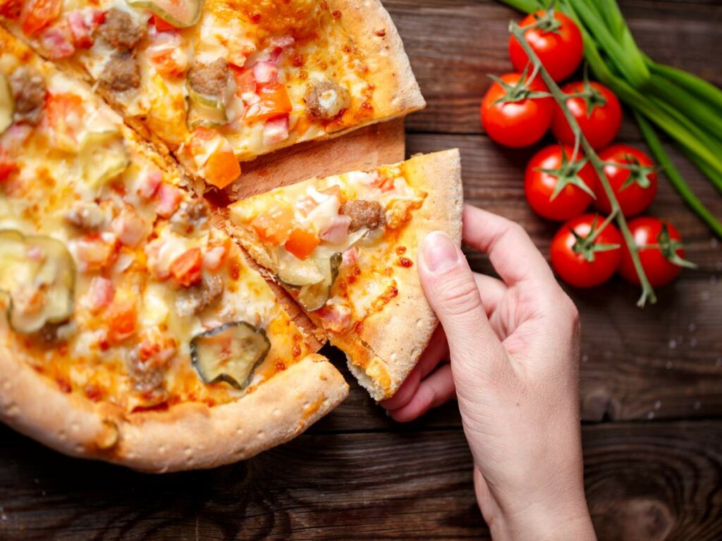 A hand picks up a slice of pizza topped with melted cheese, tomatoes, pickles, and meat. Fresh tomatoes and green onions are on the wooden table beside the pizza.