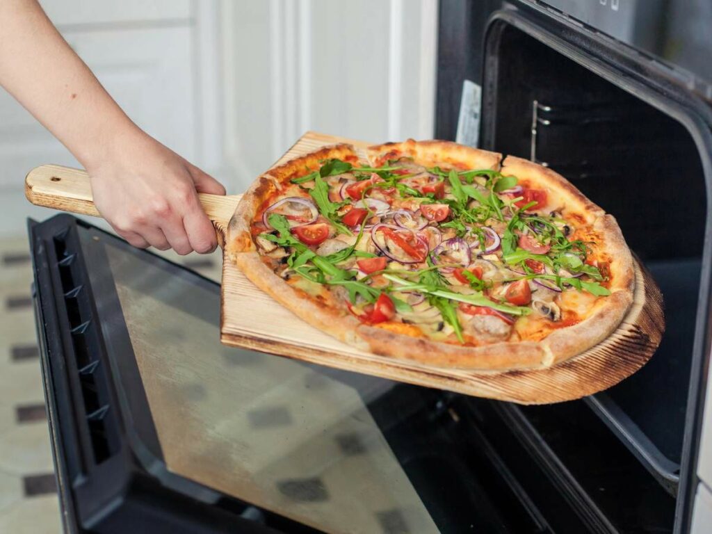 A person is using a wooden pizza peel to place a homemade pizza, topped with vegetables and greens, into an oven.