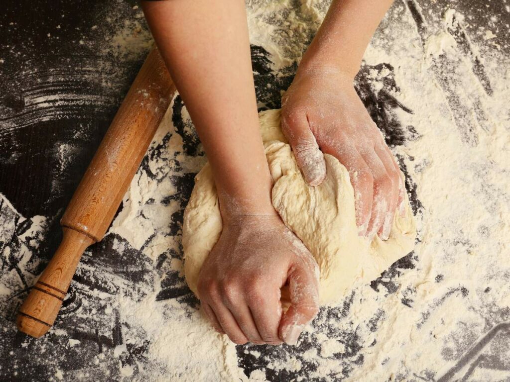 Hands knead dough on a floured surface with a wooden rolling pin nearby.