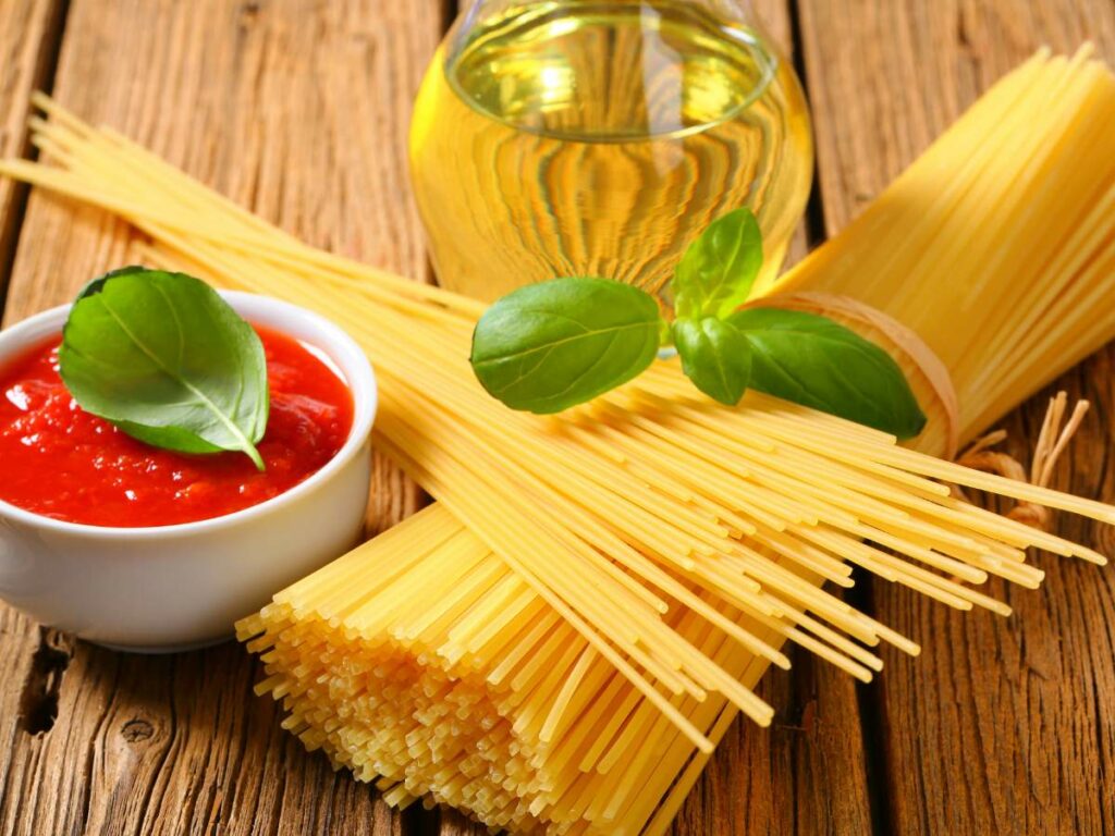 Uncooked spaghetti, a small bowl of tomato sauce, and a glass jug of oil set on a wooden surface.