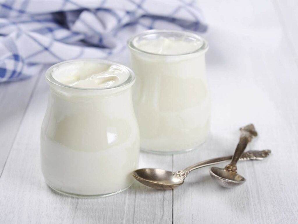 Two glass jars filled with yogurt sit on a white wooden surface, accompanied by two silver spoons and a blue checkered cloth in the background.