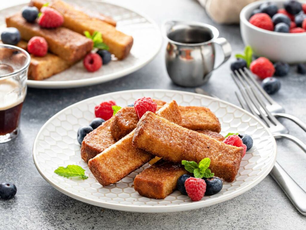 A plate of French toast sticks garnished with a bowl of berries and a small pitcher in the background.