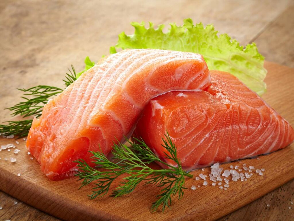 Two raw salmon fillets with salt on a wooden cutting board, garnished with a leaf of lettuce.