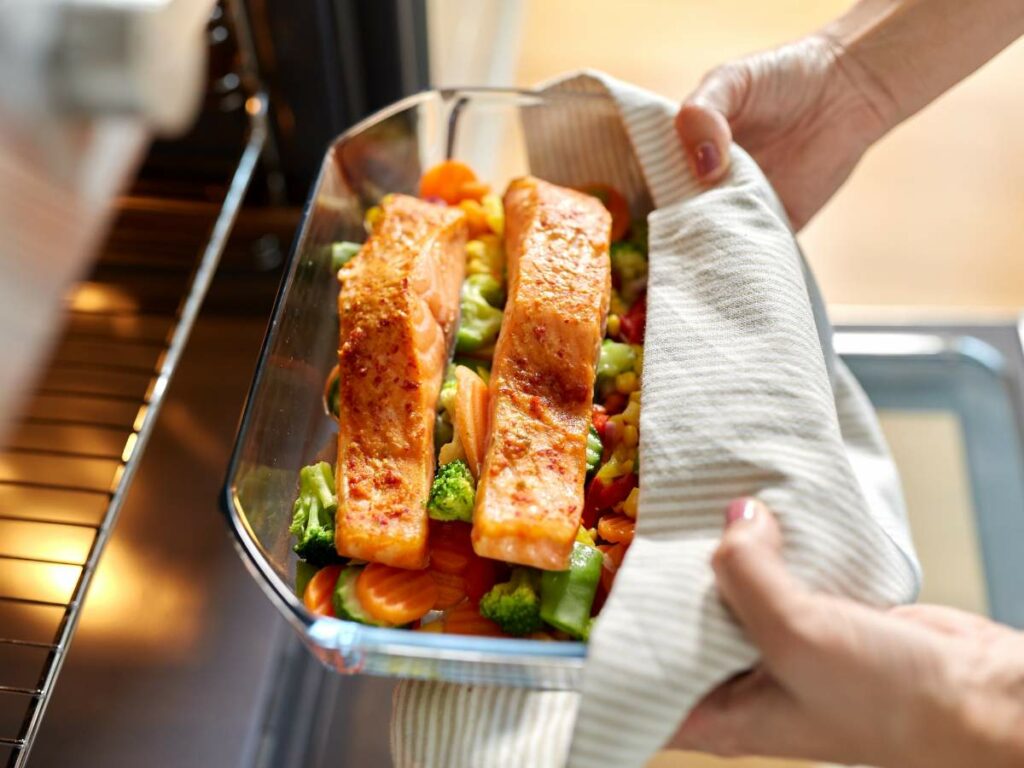 Hands holding a glass baking dish with two salmon fillets on top of mixed vegetables, about to be placed into an oven.