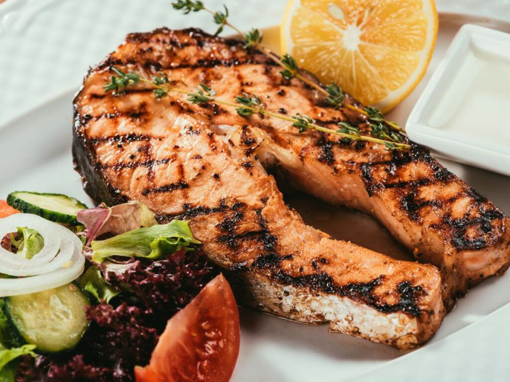 A grilled salmon steak topped with herbs, served on a white plate with a side salad and lemon wedge.