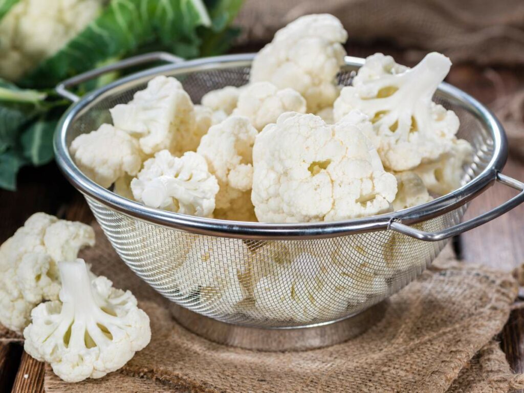 A colander filled with fresh cauliflower florets sits on a burlap cloth.