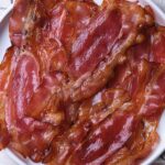 A plate of crispy maple bacon strips arranged on a white dish atop a checkered cloth.