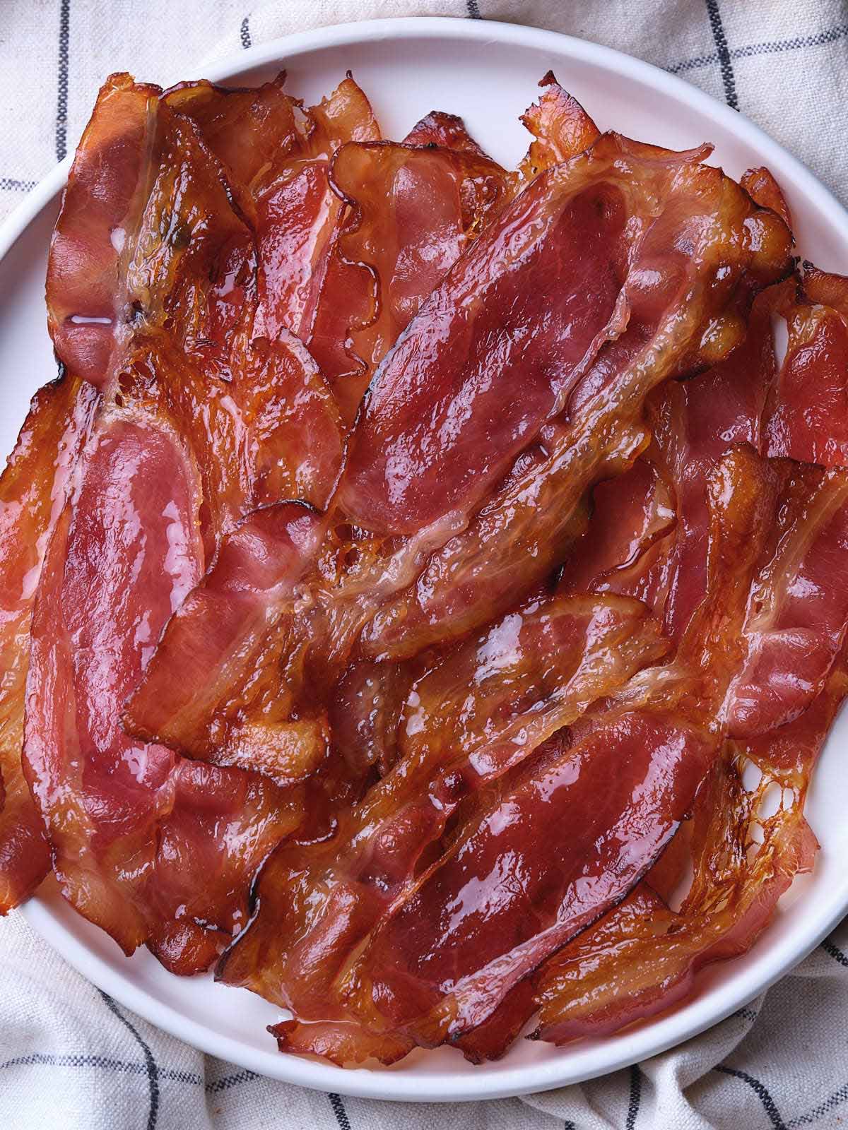 A plate of crispy maple bacon strips arranged on a white dish atop a checkered cloth.