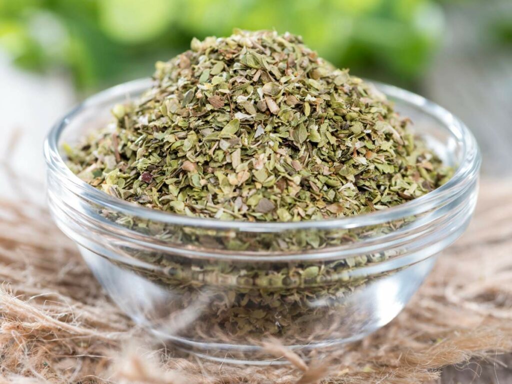 A small glass bowl filled with dried oregano leaves sits on a textured surface.