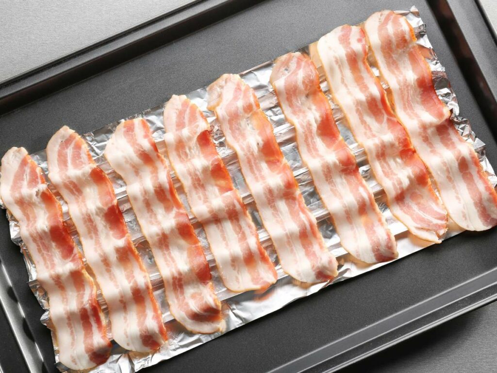 Uncooked strips of bacon arranged side by side on a baking sheet lined with aluminum foil.