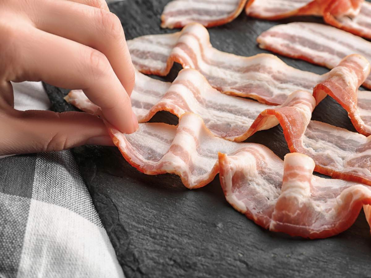 A hand arranging uncooked bacon strips on a black slate surface.