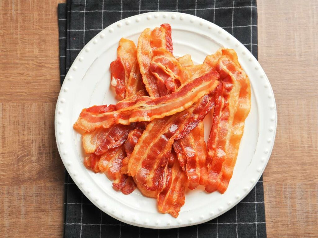 A plate of cooked bacon strips on a white plate with a black and white checkered napkin underneath.