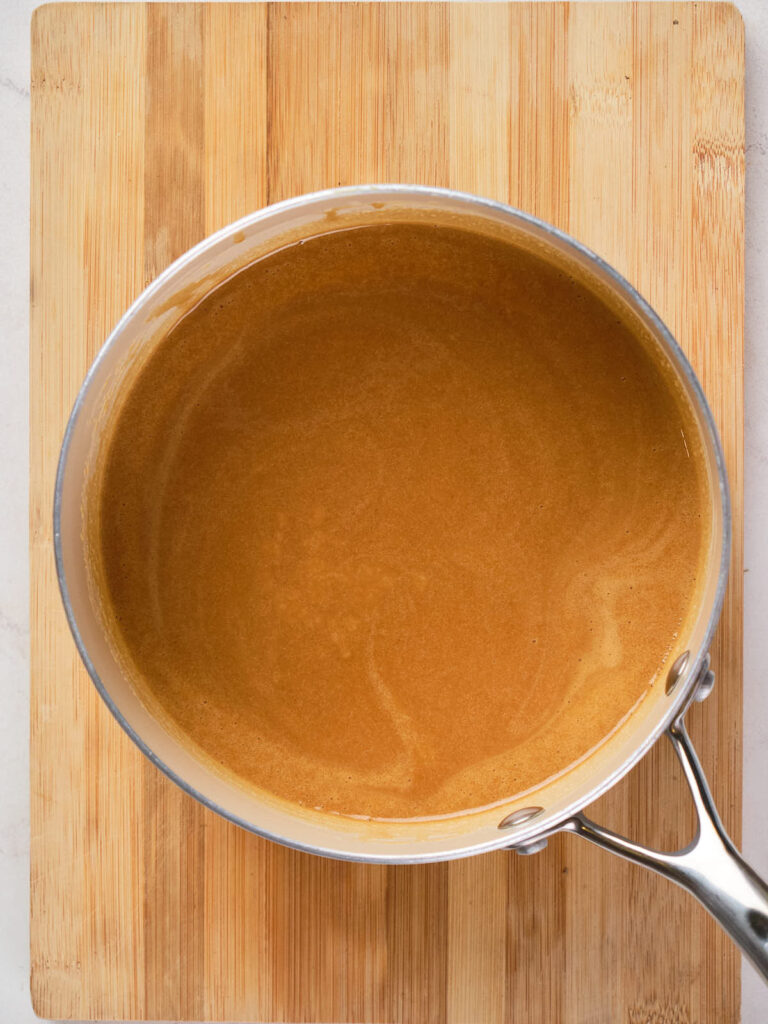 A saucepan containing a melted peanut butter mixture sits on a wooden cutting board.