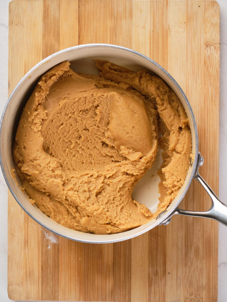 A pot filled with thick, smooth peanut butter dough or batter, placed on a wooden cutting board.